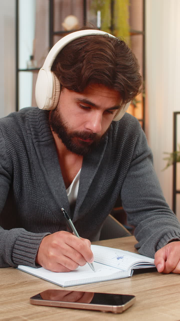 Indian man listens to audiobook on headphones takes notes in notebook focused intense study session