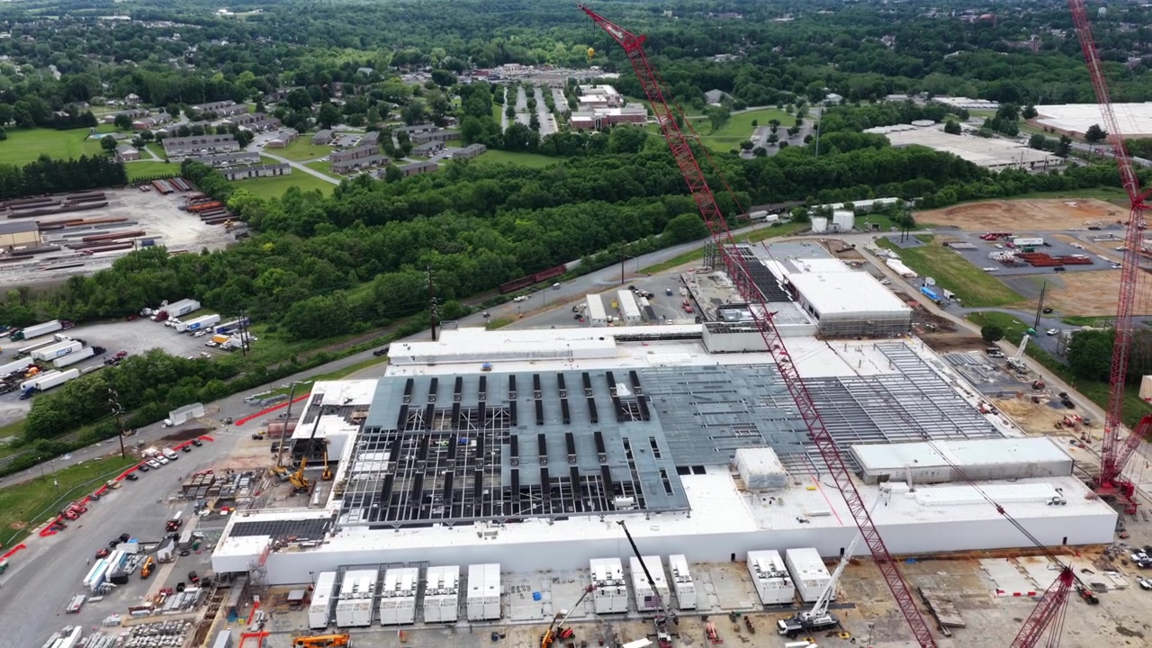 Imponenti gru rosse si ergono sopra il tetto incompiuto di un data center, rimorchi da costruzione e lotti di terra si estendono su un paesaggio suburbano boscoso. Lancaster, Pennsylvania, USA. Ripresa aerea all'indietro con drone.