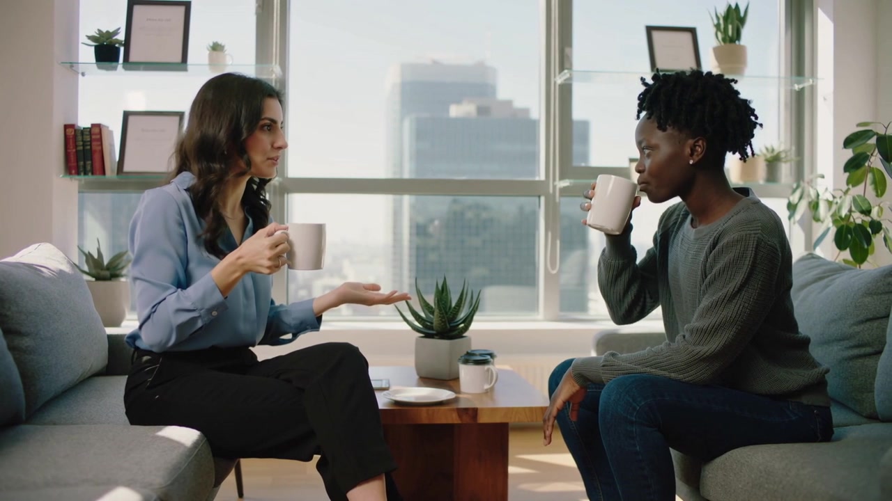 Finishing sip, left woman in blue blouse speaking, gesturing, emphasizing in lounge, holding mug