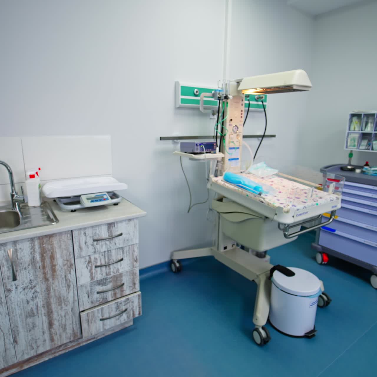 Little bed with equipment for reanimation of newly born children. Sink with table and weighs on it on the left. Stall with diapers and other necessities on the right