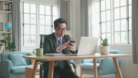 asian businessman wearing glasses and business suit using mobile phone while using the computer for working at home.