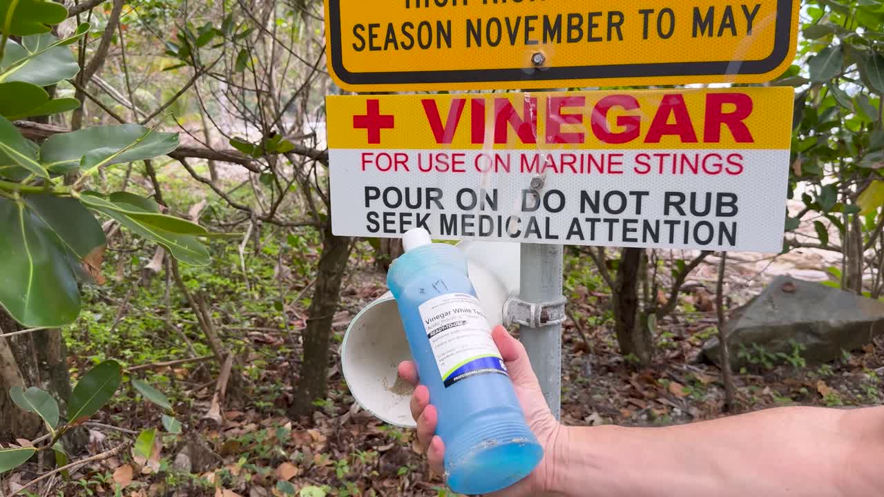A person retrieves vinegar from a dispenser near a marine stinger warning sign in a tropical setting