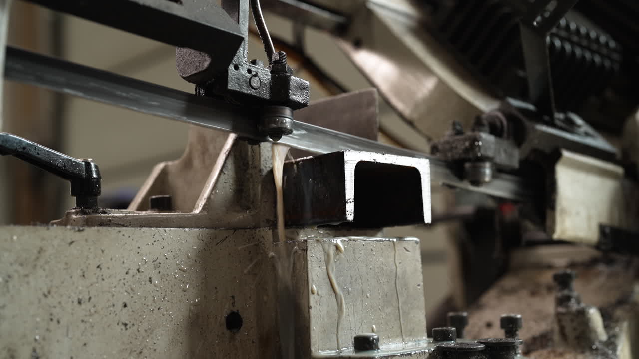 Metal cutting bandsaw slicing through a steel beam in an industrial workshop, coolant dripping on blade