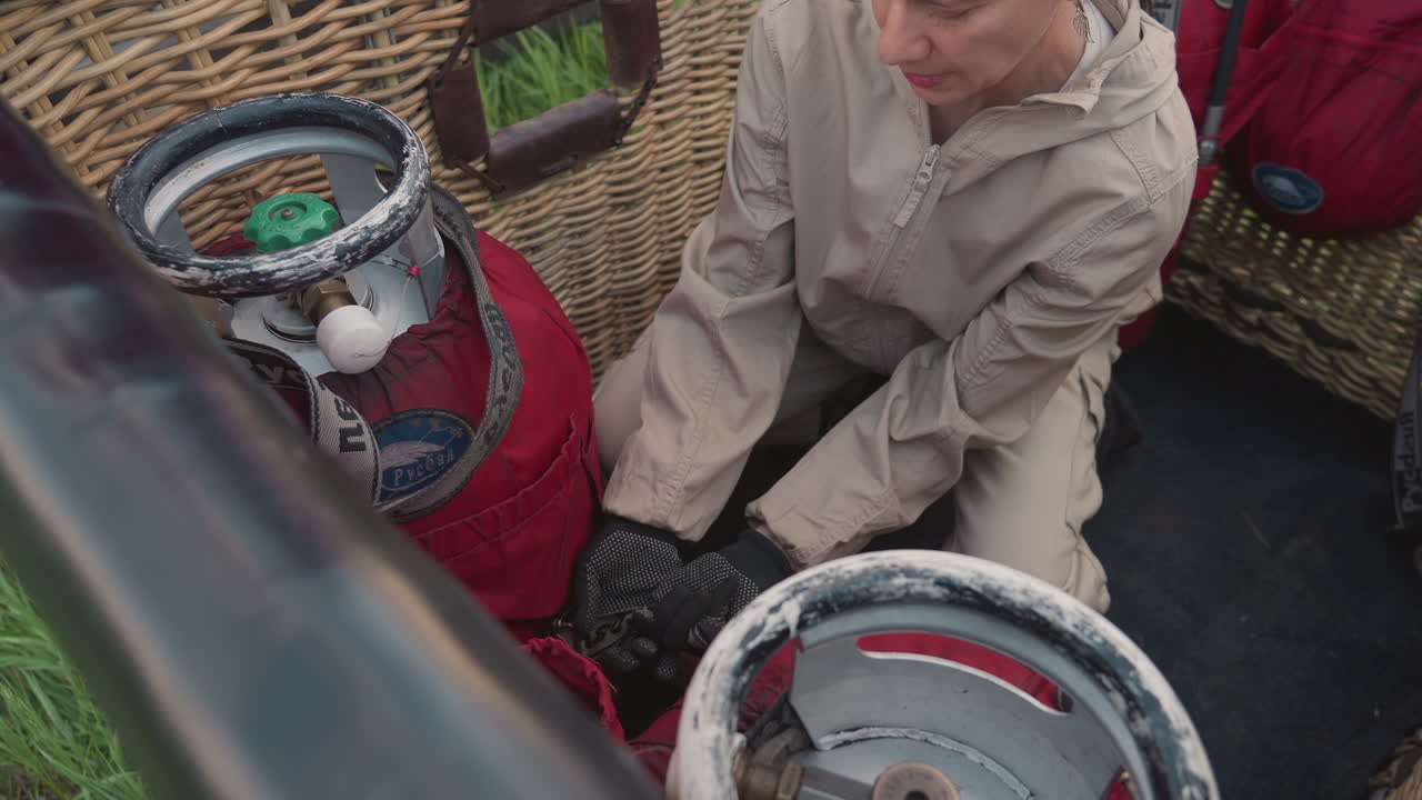 woman fastening gas cylinder in hot air balloon wicker basket with safety straps and gloves while crew prepares colorful envelope and equipment on grassy field under cloudy sunset sky