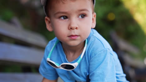 charming baby boy in cap, blue t-shirt and with sunglasses is on the bench outdoors. cheerful kid stands up and walks away. close up. blurred backdrop.