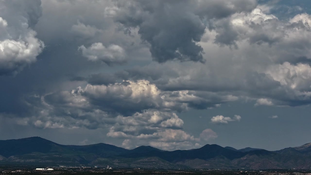 4K UHD drone-timelapse af et komplekst vejrsystem. Tunge, flerlagede stormskyer dannes og skifter dynamisk over en fjern, silhuetformet bjergkæde og en udstrakt højtliggende ørkendal.