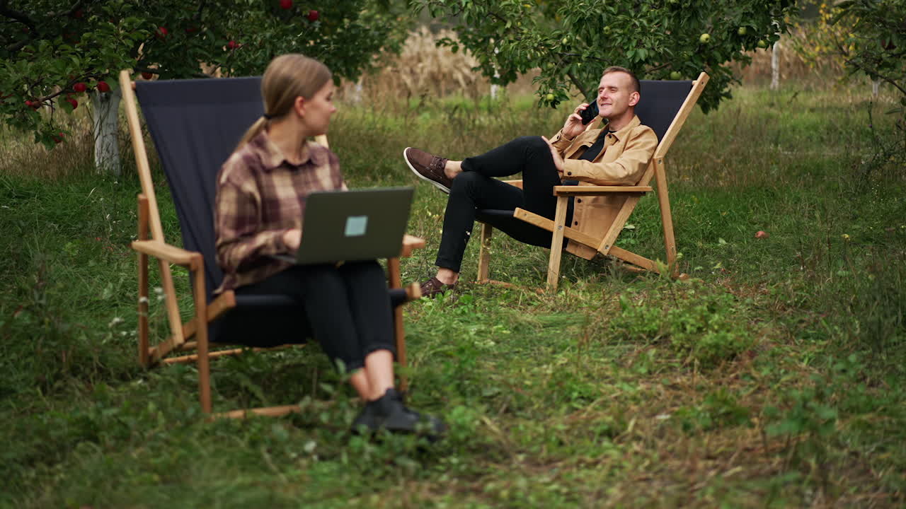 Married couple working online in their garden. Woman works on laptop and man speaking on the phone. Family freelancers working remotely.