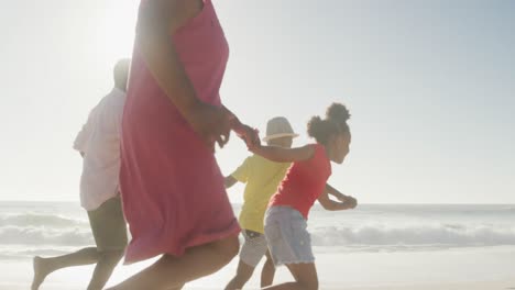 Smiling-senior-african-american-couple-running-with-grandchildren-on-sunny-beach