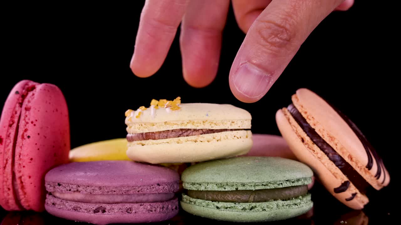 A hand reaches in to pick up a yellow macaron from a neat row of assorted colorful macarons on a glossy black surface under bright studio lighting