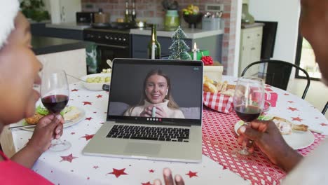 african american couple with wine using laptop for christmas video call with happy woman on screen