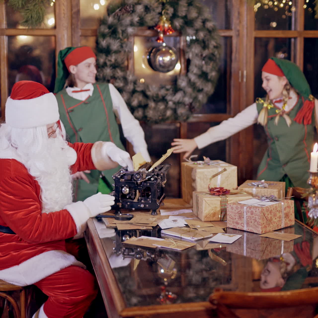 Merry atmosphere with elves and Santa Claus. Santa with white beard in red costume is typing letter on the background of funny elves. New Year's decorations in Santa's residence.