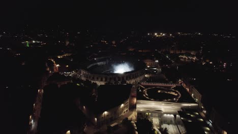 Rotación-De-Drones-En-Las-Arenas-De-Nîmes-En-Medio-De-La-Noche,-La-Gente-Está-Viendo-El-Concierto-Y-Hay-Luces-De-Varios-Colores