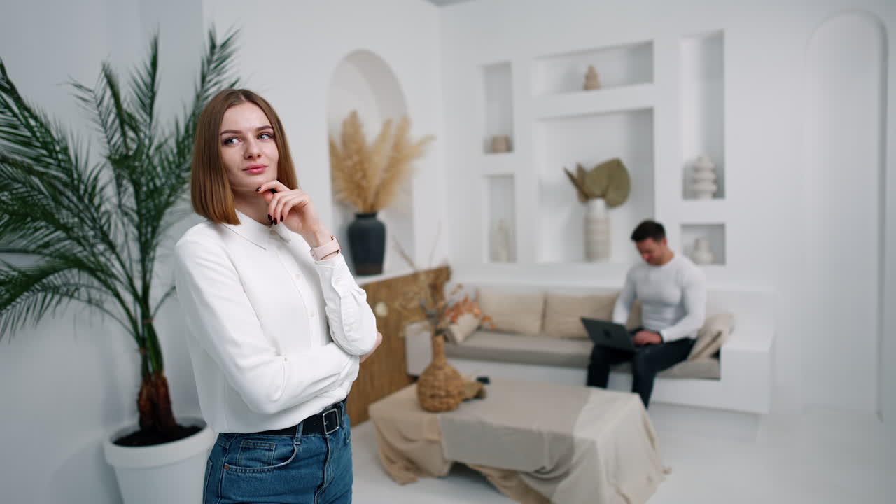 Confident pensive young woman stands in the cozy room. Man sitting on sofa works on laptop. Blurred backdrop.
