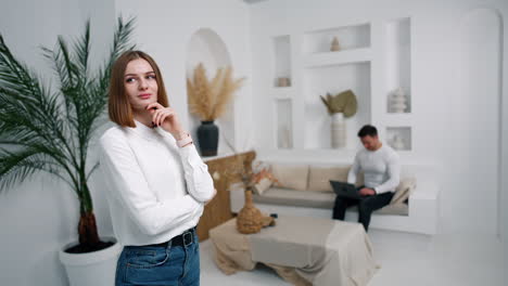 confident pensive young woman stands in the cozy room. man sitting on sofa works on laptop. blurred backdrop.
