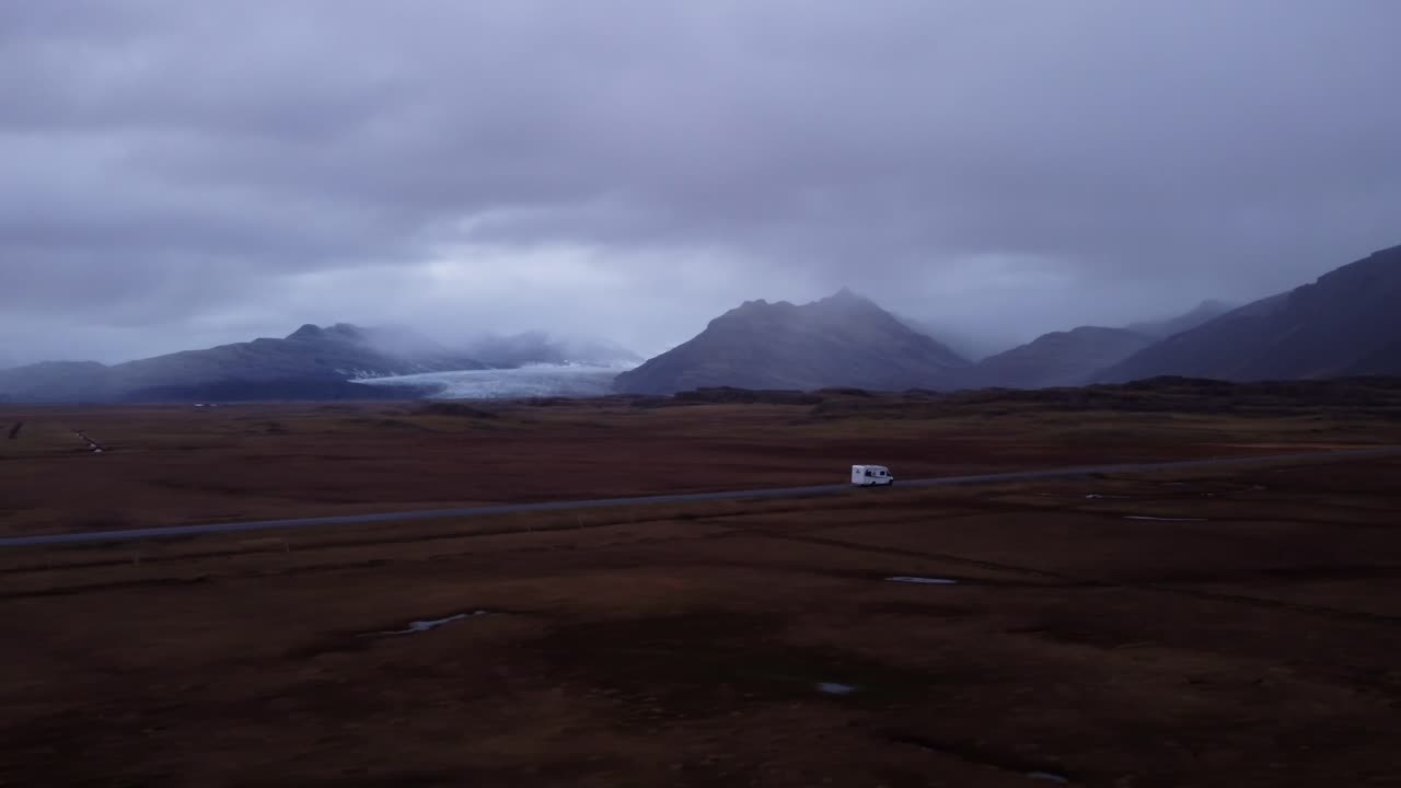 An expansive aerial shot captures a motorhome traveling along a road that cuts through a vast, rugged, and open Icelandic landscape