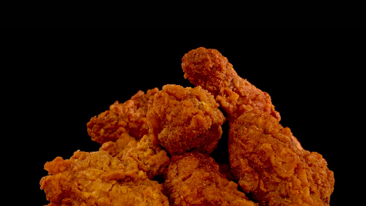 Golden, crispy fried chicken pieces are slowly zoomed in on against a black background, highlighting spicy seasoning and crunchy texture under even studio lighting