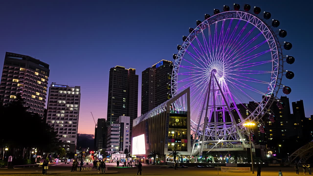 Vibrant Night Sokcho Cityscape With Majestic Sokcho Eye Ferris Wheel Illuminated. Street is bustling with people activity walking along the sidewalks. Skyscrapers and buildings towers in background