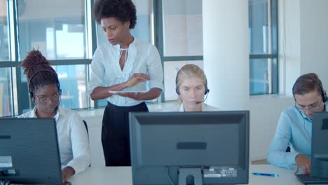 Serious-female-call-center-leader-helping-employee-in-headset