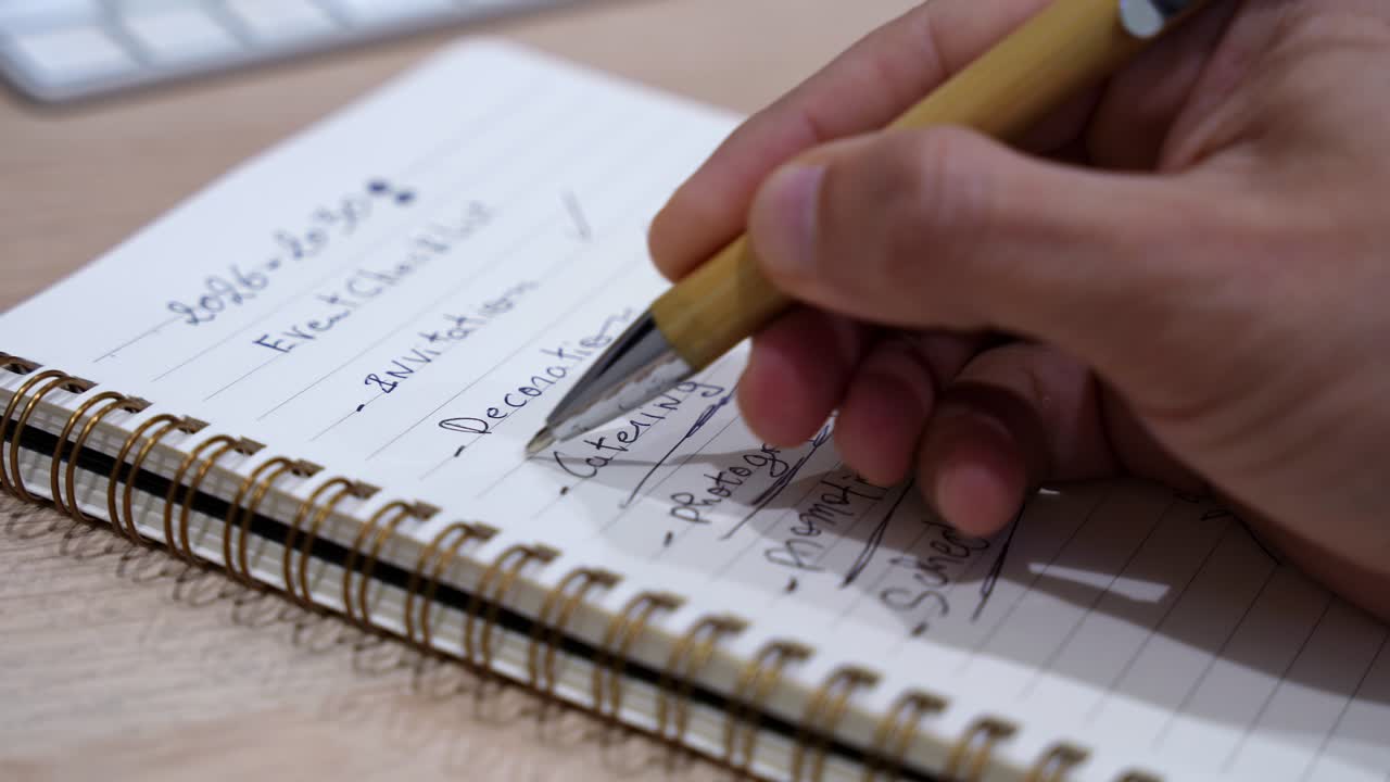 Close-up of a hand writing a job task checklist in a spiral notebook on a wooden desk indoors, with pen tip in focus and soft office background blur