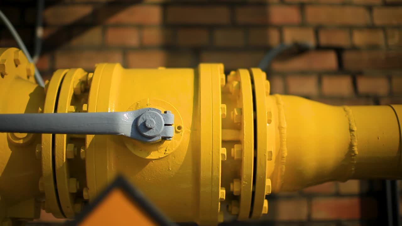 Primo piano di un gasdotto giallo brillante con flange, sullo sfondo di un muro di mattoni, che evidenzia l'infrastruttura industriale.