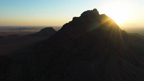 Aerial-view-around-a-rocky-mountain-in-the-Namib-desert-with-a-sunburst-in-Namibia