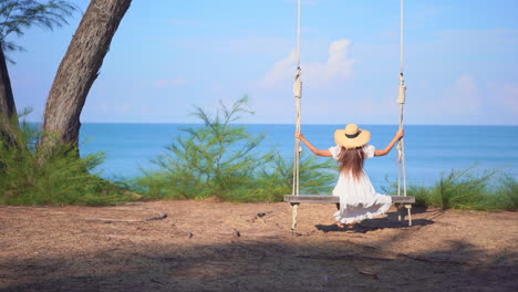 Vestida-Con-Un-Clásico-Vestido-De-Verano,-Una-Mujer-De-Espaldas-A-La-Cámara-Se-Balancea-Suavemente-En-Un-Gran-Columpio-De-Madera-Mirando-El-Horizonte-Del-Océano