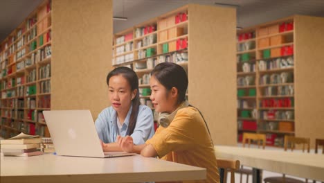 asian woman student teaching her classmate on a laptop while sitting on a table in the library