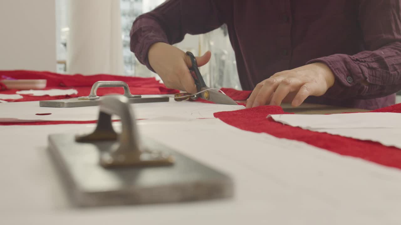 A woman cuts fabric along paper outlines, using a heavy iron weight to secure the layers while she trims the shapes with scissors