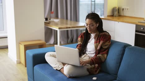chica positiva con rastas sentada en el sofá con una computadora portátil en las rodillas