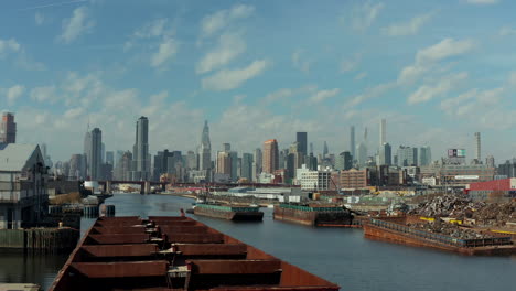 Barges-for-transporting-material-on-water-in-industrial-borough.-Modern-downtown-skyscrapers-in-background.-New-York-City,-USA