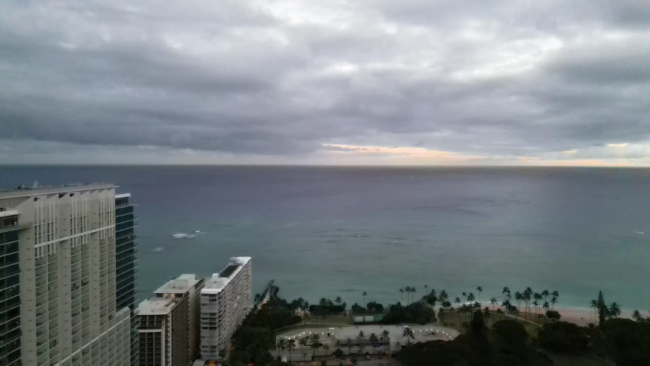 Vista aerea da un grattacielo dell'oceano, degli hotel costieri e delle palme sulla spiaggia di Waikiki sotto un cielo drammatico e nuvoloso prima di una tempesta, con un accenno di tramonto all'orizzonte