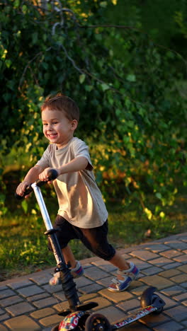 little toddler boy approaching camera riding on his scooter. adorable smiling happy kid having fun active time in the park at sunset. vertical video.