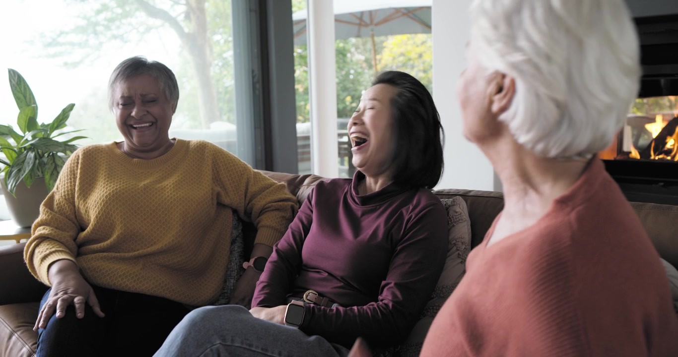 Diverse female friends turning toward right senior woman telling story on sofa by fireplace smiling