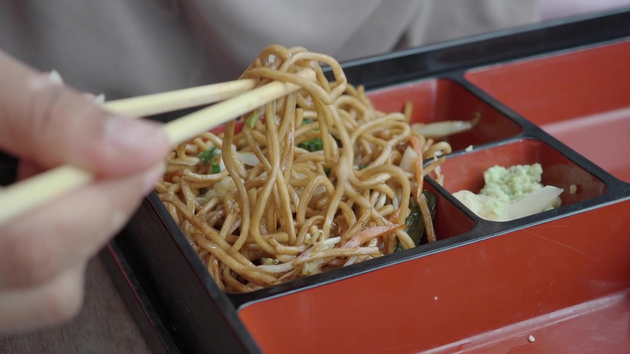 Pessoa comendo macarrão salteado com pauzinhos de uma caixa bento.