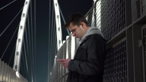 Junger-Männlicher-Teenager,-Der-Nachts-An-Seinem-Telefon-Auf-Einer-Fußgängerbrücke-Steht