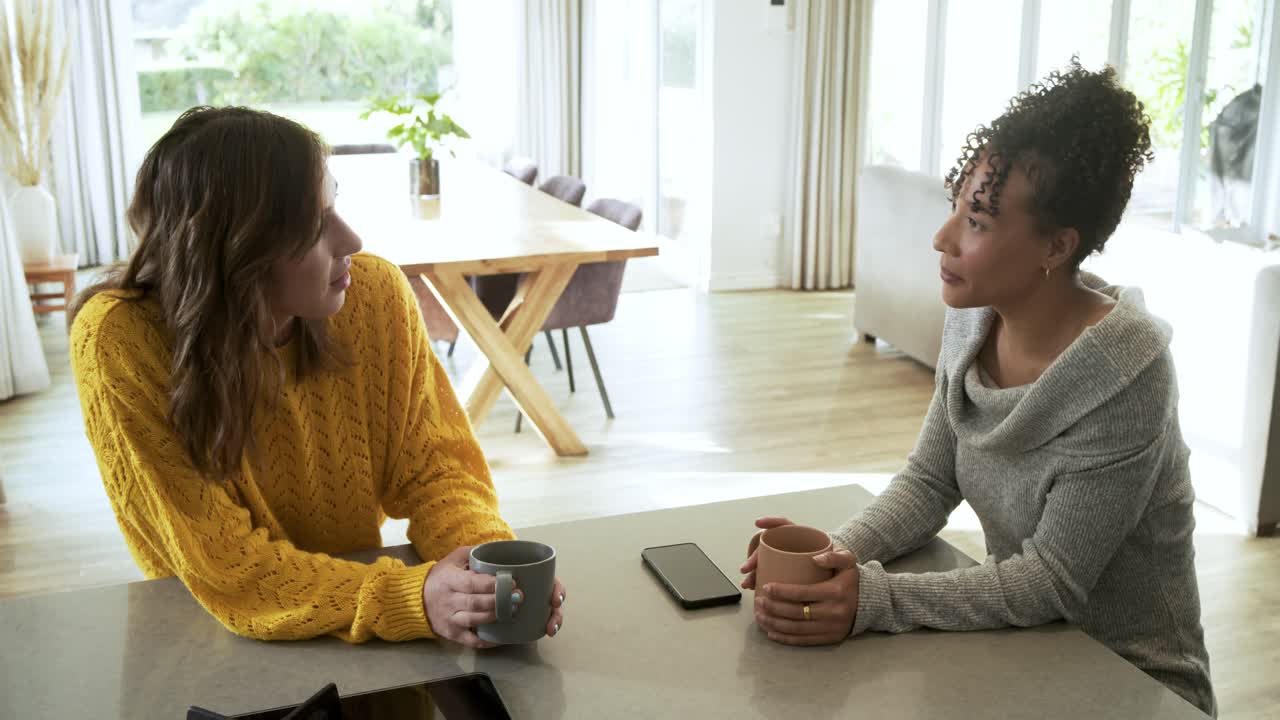 Amigas diversas conversando enquanto mulher de suéter mostarda se inclina segurando canecas na ilha da cozinha
