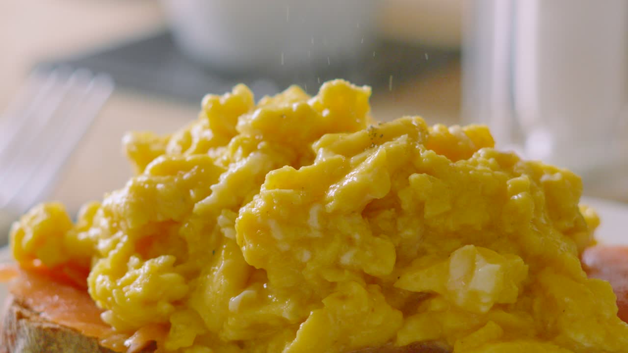 Close Up of Fine Pepper Granules Falling on Delicious Morning Breakfast Scrambled Eggs on Smoked Salmon with Coffee Cups and Fork in Background. Slow Motion Food Photography.