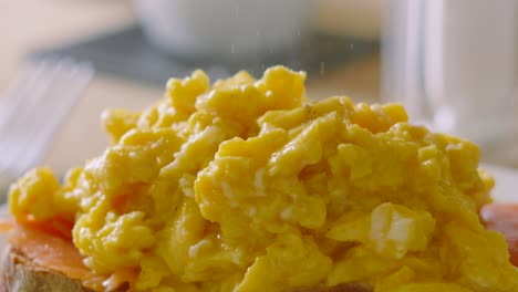 close up of fine pepper granules falling on delicious morning breakfast scrambled eggs on smoked salmon with coffee cups and fork in background. slow motion food photography.