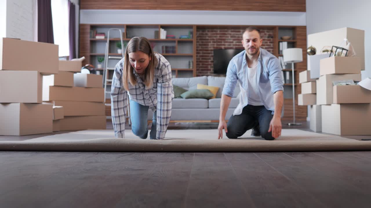 Young couple unrolling the carpet in new apartment on moving day. New accommodation or rented flat. Happy homeowners on relocation day. Real estate and tenancy concept. Loan mortgage for young family