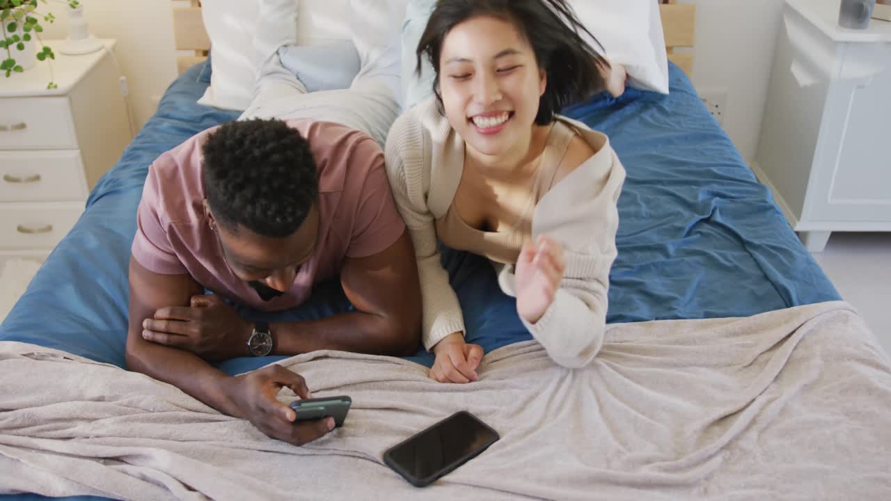 Felice coppia multietnica che usa lo smartphone e sdraiata in camera da letto