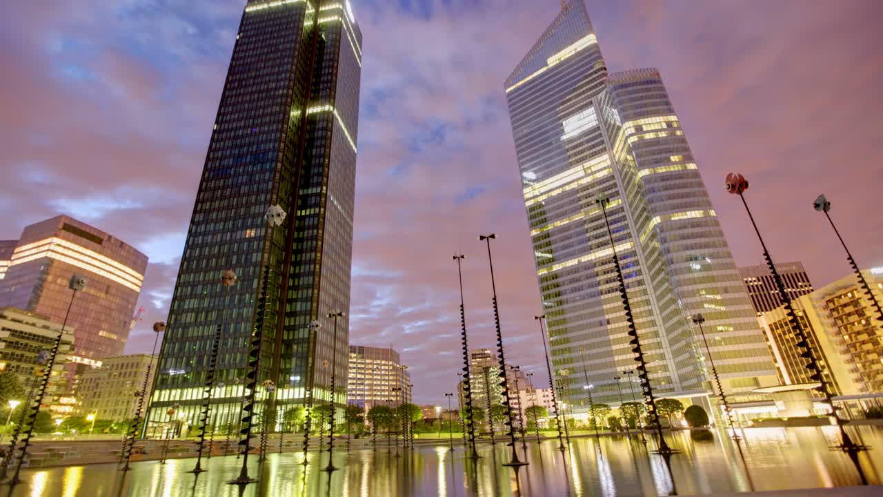 el timelapse captura un sueño del horizonte de la ciudad por la noche desde un ángulo bajo, altos edificios modernos y farolas se deleitan en el reflejo de las luces de la ciudad contra un cielo nublado naranja