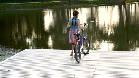 a little girl with pigtails down with a bicycle to the pond in the city park at sunset. she stands on a wooden bridge and looks at the water.