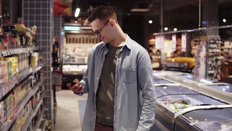 a good looking young caucasian guy in glasses and blue shirt doing shopping in the supermarket, walking by aisle and stops in front the shelf. makes a photo with smartphone, looking for special offers or discount