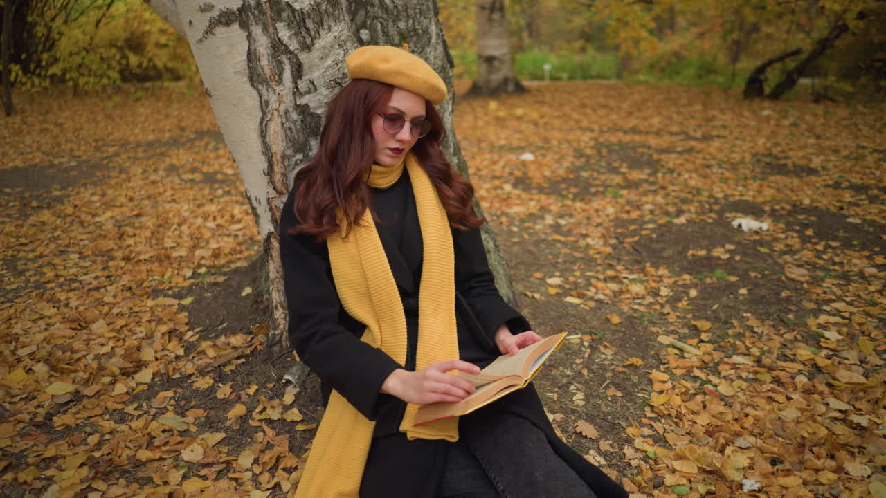 Туристка в уединении задумчиво листает страницы книги, сидя в одиночестве на природе, одетая в желтый берет и шарф, окруженная осенними листьями, погруженная в мирный момент чтения.