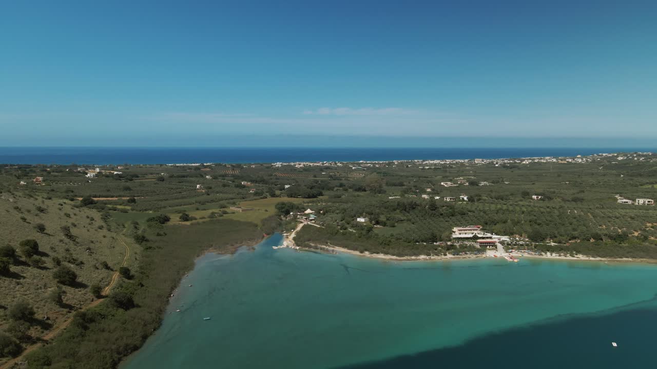 Turquoise waters of Lake Kournas meet the shoreline near a small coastal village with distant view of the sea and Cretan countryside under a bright blue sky