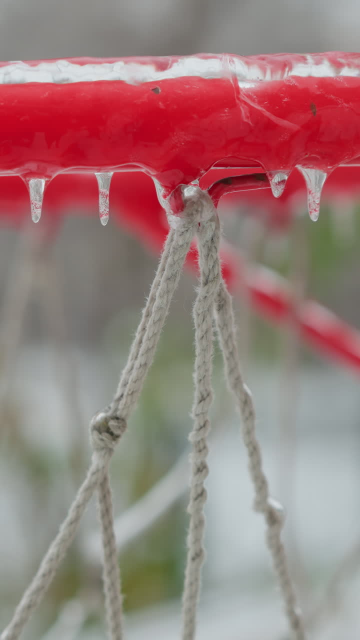 Vivid red basketball rim covered in ice with hanging icicles, close-up, against a blurred snowy playground, illustrating winter's effect on outdoor sports facilities