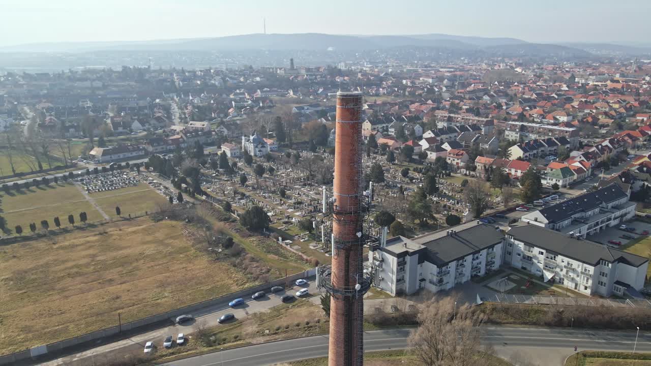 Flying around a tall brick industrial chimney surrounded by a residential neighborhood with red-roofed houses. Drone view of the cityscape.