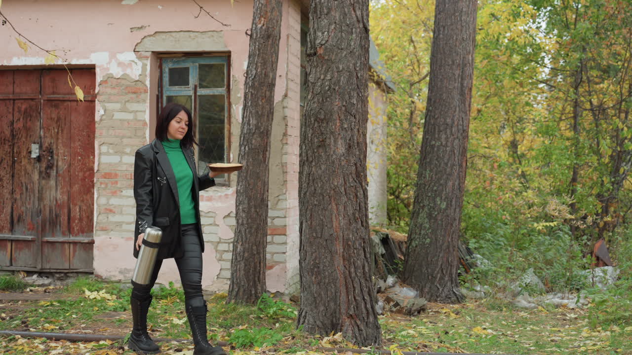 rockera con chaqueta de cuero en el exterior de una cabaña, fondo de bosque otoñal con hojas caídas y pinos altos, fachada rosa desgastada y puerta rústica, pose segura con termo, caminando cerca de troncos de árboles