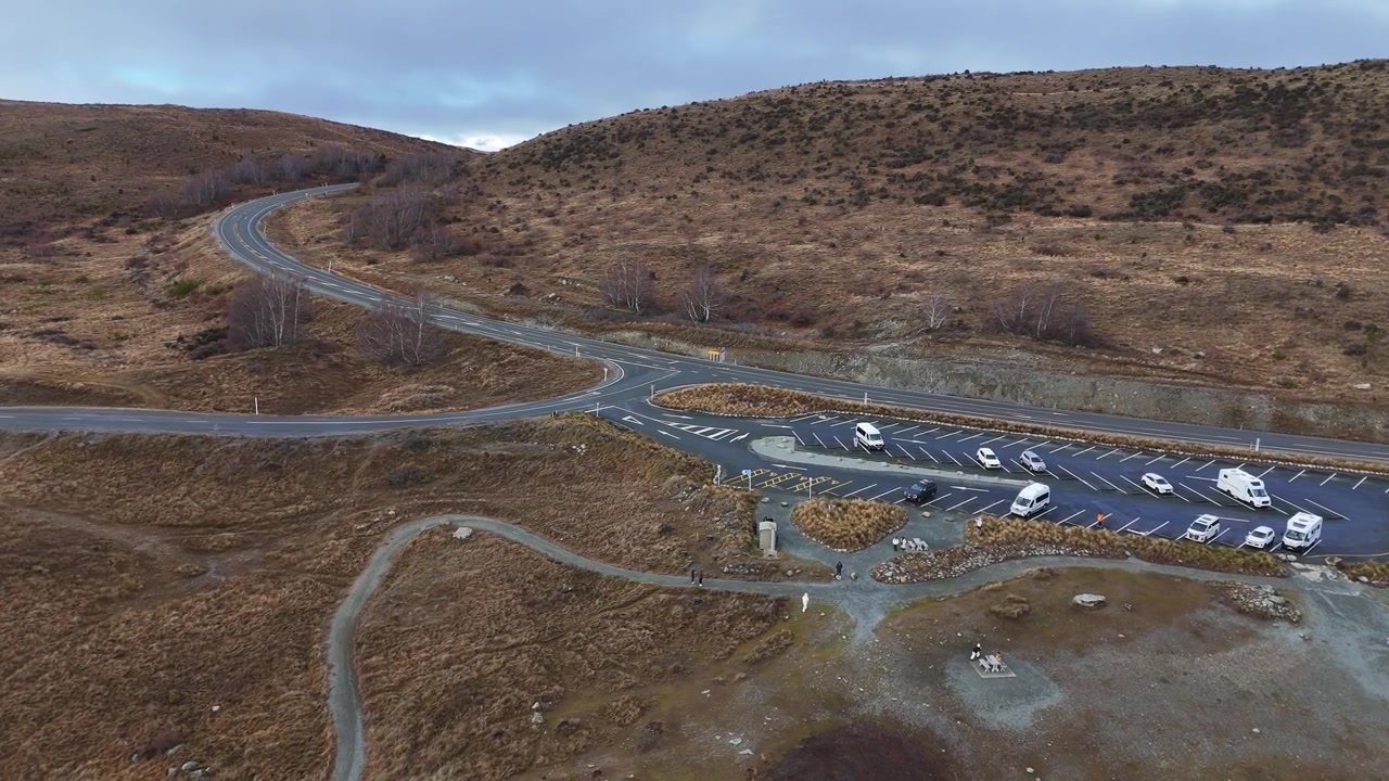 Yeni Zelanda'da Pukaki Gölü ziyaretçi otoparkı ve çevresindeki yolların üzerinden sağa doğru kayan hava drone çekimi, engebeli yüksek araziyi ve dramatik alp manzarasını gözler önüne seriyor.