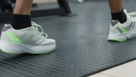 leg view of athlete wearing white neon green sneakers and black socks walking on textured gym mat past blurred weight bench and machine in bright fitness studio corridor